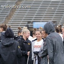 A group of people, likely athletes, are gathered together on a sports field. They are wearing black jackets with some individuals dressed in white sports uniforms underneath. A few people have their hair tied up in buns, and most are engaged in conversation. The background shows tiered stadium seats, suggesting the setting is an outdoor sports venue.